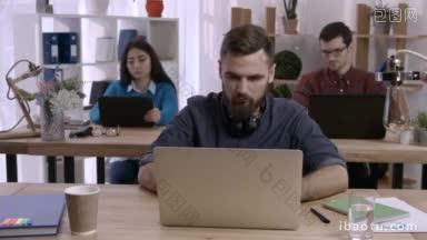 疲惫的潮人商人与胡子伸展手臂在笔记本电脑前，而坐在办公桌在现代办公室自由职业者